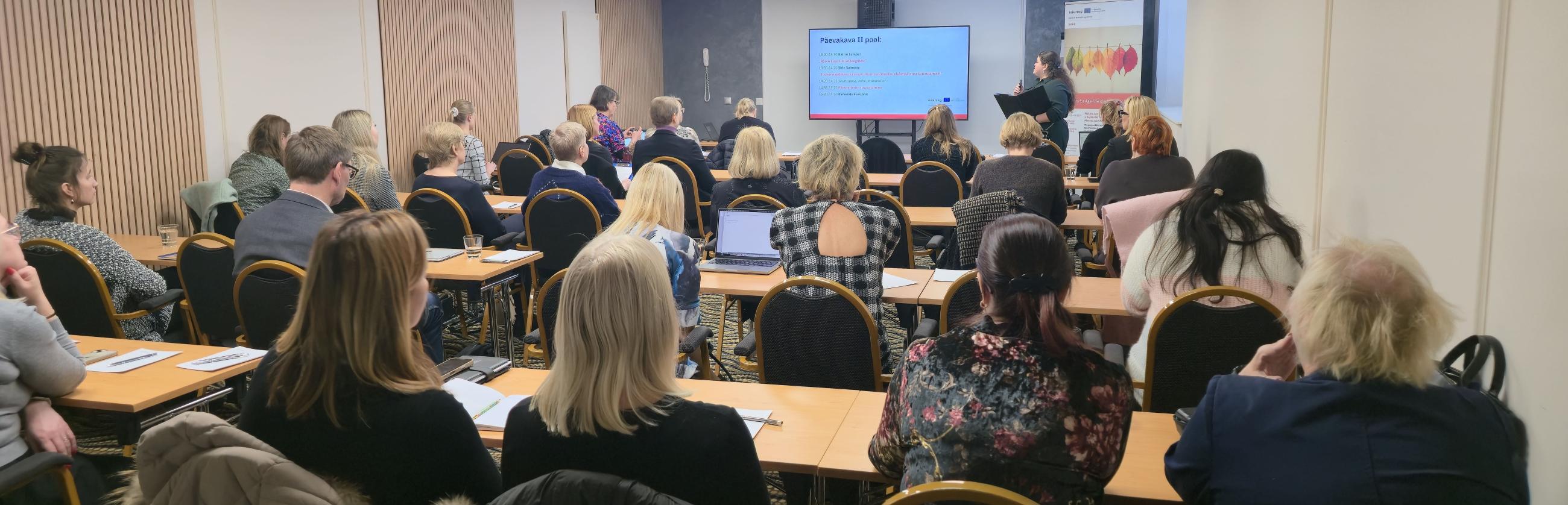 Fotol on konverentsiruumis toimuv koolitus või seminar. Ruumi eesosas seisab esineja, kes tutvustab ekraanil kuvatavat esitlust, samal ajal kui kuulajad istuvad laudade taga ja jälgivad ettekannet. Osalejaid on palju ning nad on keskendunult kuulamas, mõned teevad märkmeid.