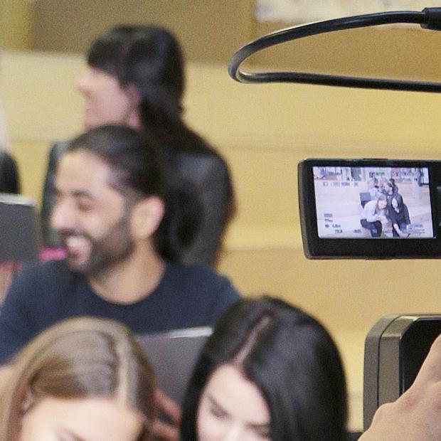 Tagaplaanil noored inimesed istumas, esiplaanil operaator kaameraga neid filmimas