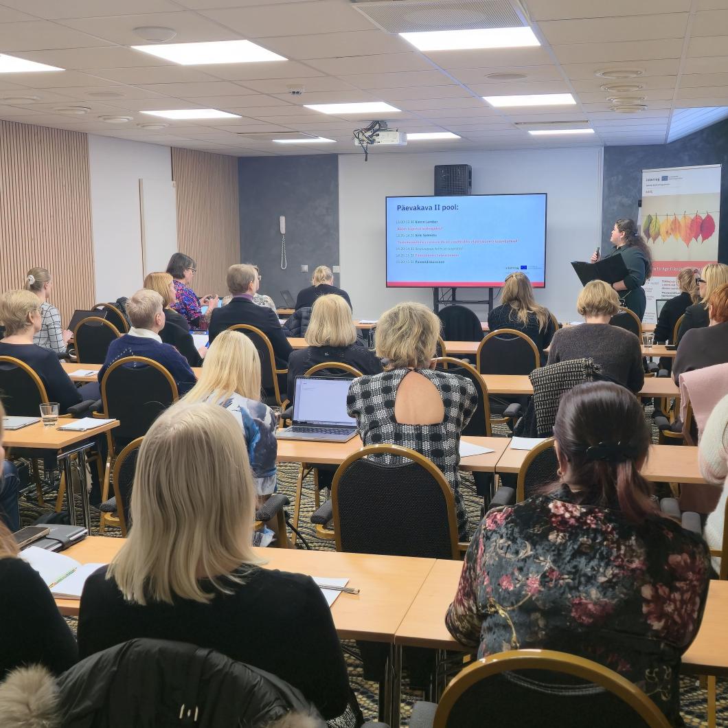 Fotol on konverentsiruumis toimuv koolitus või seminar. Ruumi eesosas seisab esineja, kes tutvustab ekraanil kuvatavat esitlust, samal ajal kui kuulajad istuvad laudade taga ja jälgivad ettekannet. Osalejaid on palju ning nad on keskendunult kuulamas, mõned teevad märkmeid.